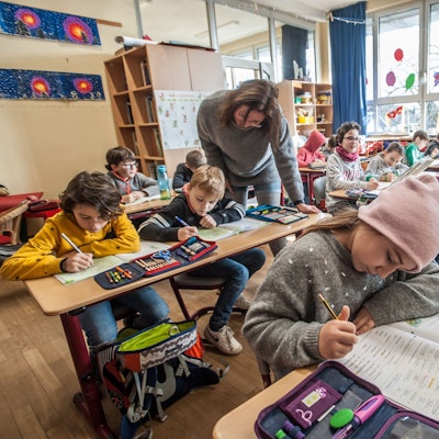 Jungen und Mädchen arbeiten in einem Klassenraum konzentriert an Aufgaben, eine Lehrerin schaut einem Jungen über die Schulter.