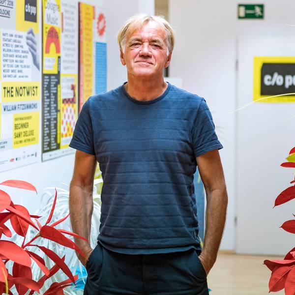 Norbert Oberhaus steht in einem Flur vor Blumen und Postern und schaut in die Kamera.