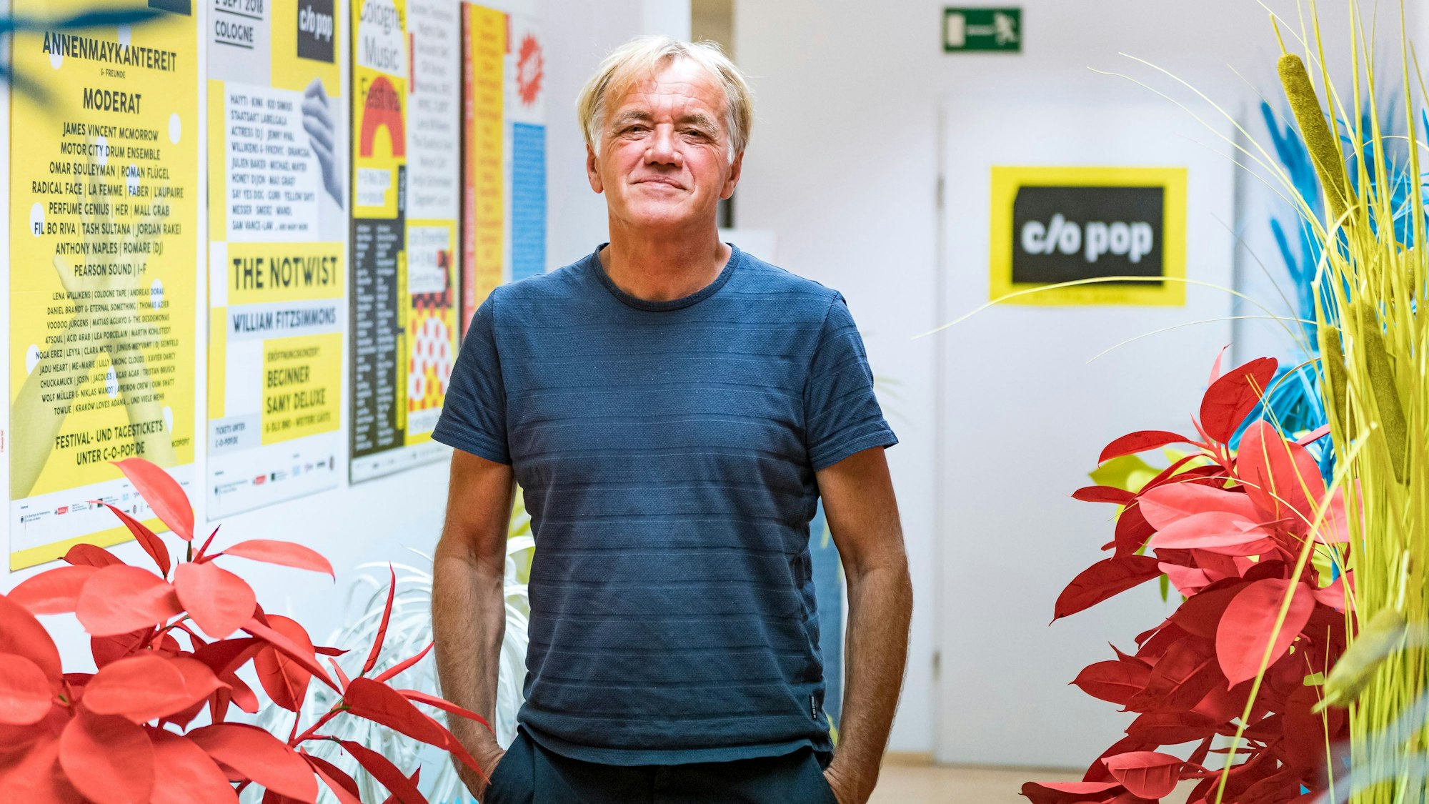 Norbert Oberhaus steht in einem Flur vor Blumen und Postern und schaut in die Kamera.