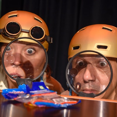 Zwei als Agenten verkleidete Schauspieler mit goldenem Helm betrachten durch eine Lupe eine Weingummi-Tüte.