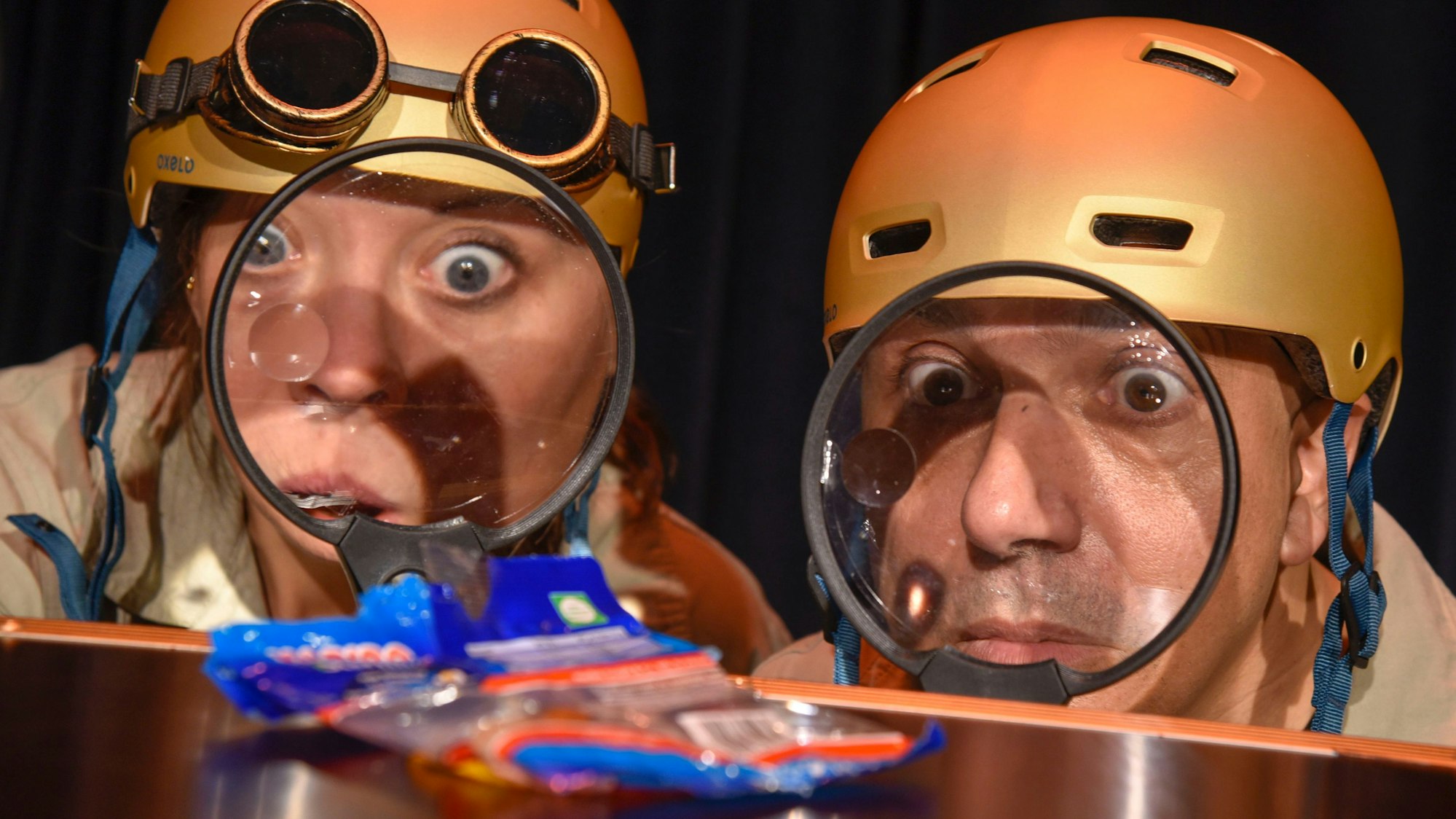 Zwei als Agenten verkleidete Schauspieler mit goldenem Helm betrachten durch eine Lupe eine Weingummi-Tüte.