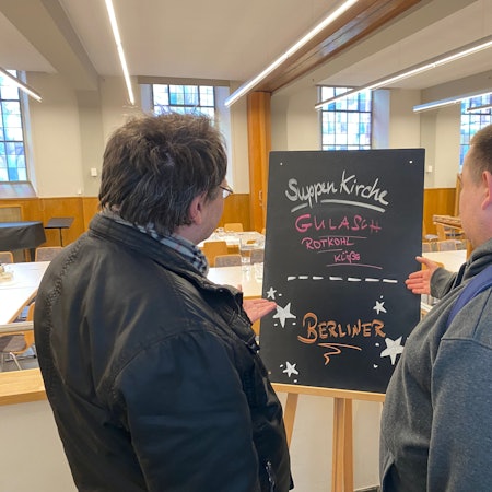 Zwei Männer stehen mit dem Rücken zur Kamera und schauen auf eine Tafel, auf der das Angebot der Suppenküche steht.