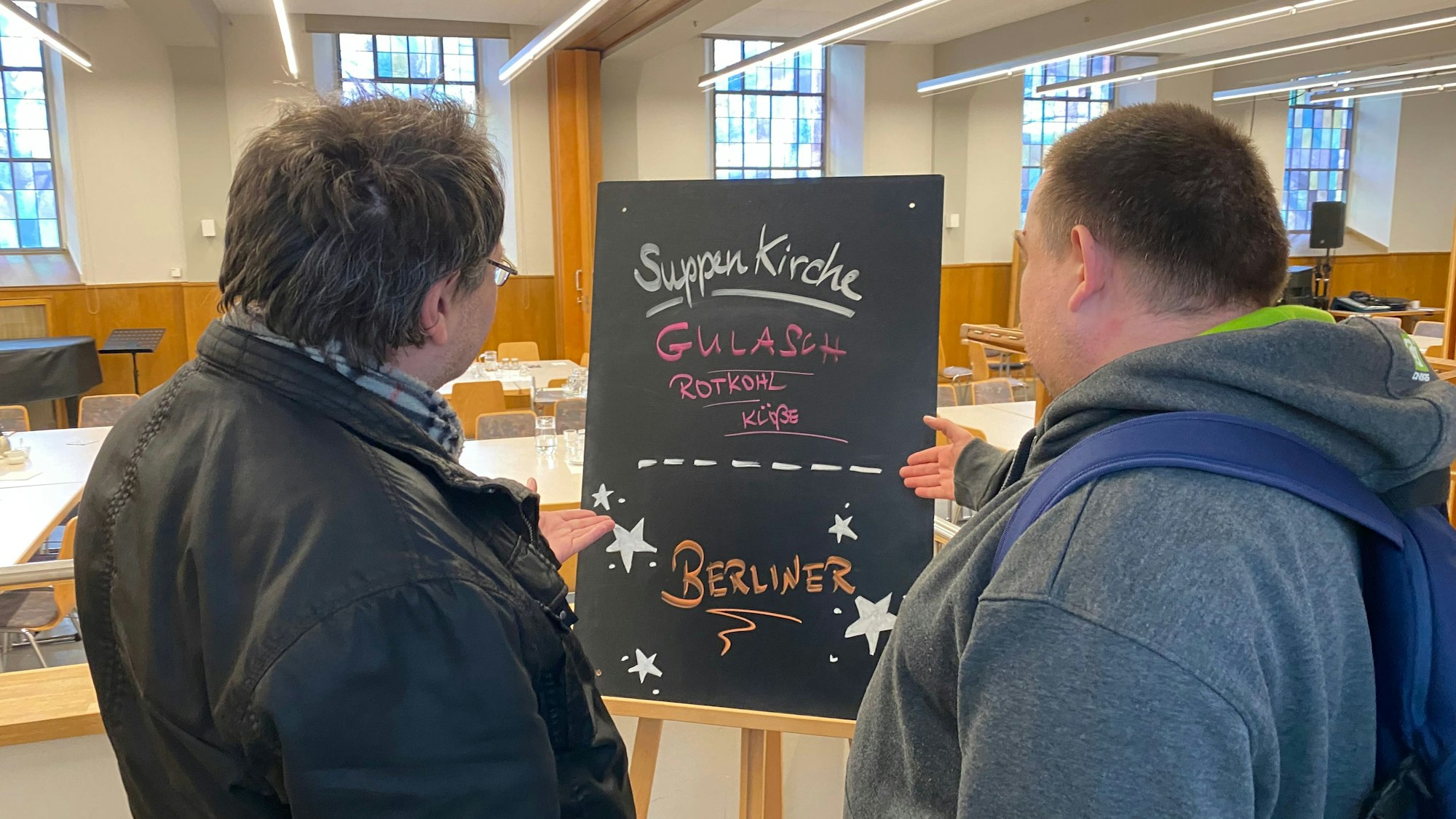 Zwei Männer stehen mit dem Rücken zur Kamera und schauen auf eine Tafel, auf der das Angebot der Suppenküche steht.