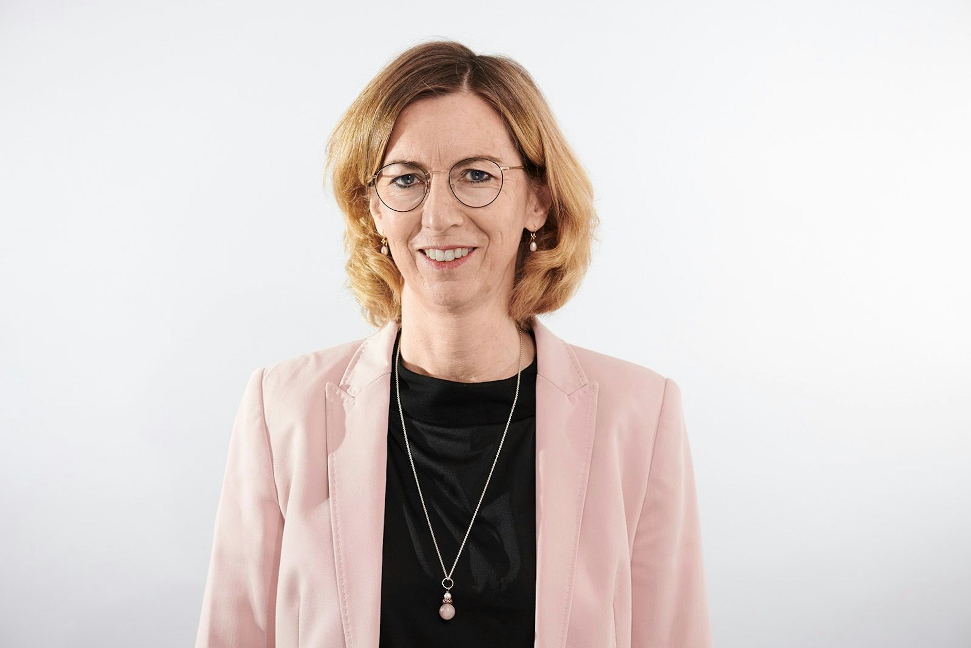 Mechthild Thamm schaut für ein Porträt in die Kamera. Sie trägt ein schwarzes Oberteil mit langer Kette, dazu einen rosafarbenen Blazer. Ihre Haare trägt sie schulterlang.