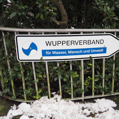 Ein Wegweiser mit der Aufschrift „Wupperverband für Wasser, Mensche und Umwelt“ vor einer grünen Hecke an einem Zaun vor der Verbandszentrale in Wuppertal.