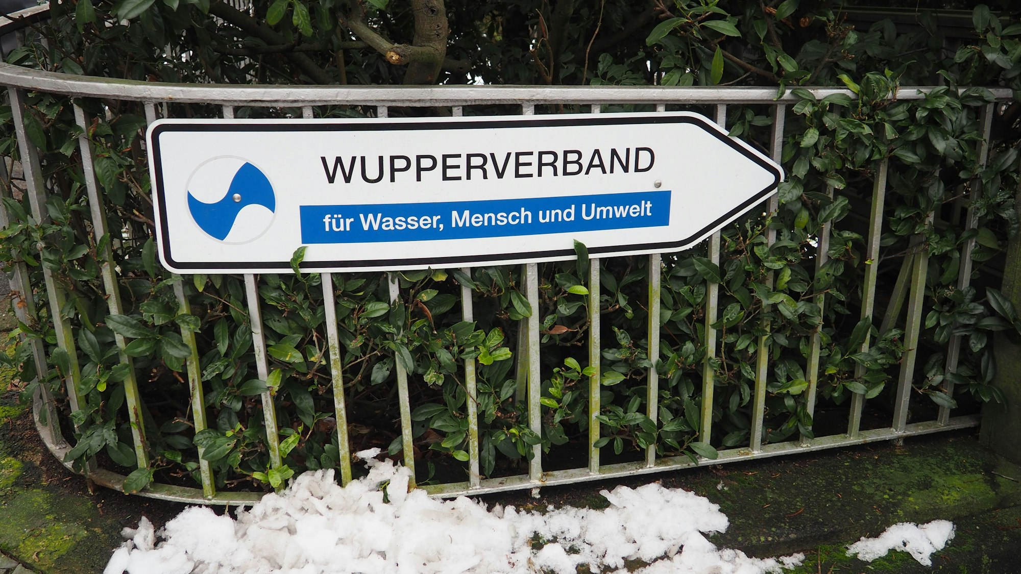 Ein Wegweiser mit der Aufschrift „Wupperverband für Wasser, Mensche und Umwelt“ vor einer grünen Hecke an einem Zaun vor der Verbandszentrale in Wuppertal.