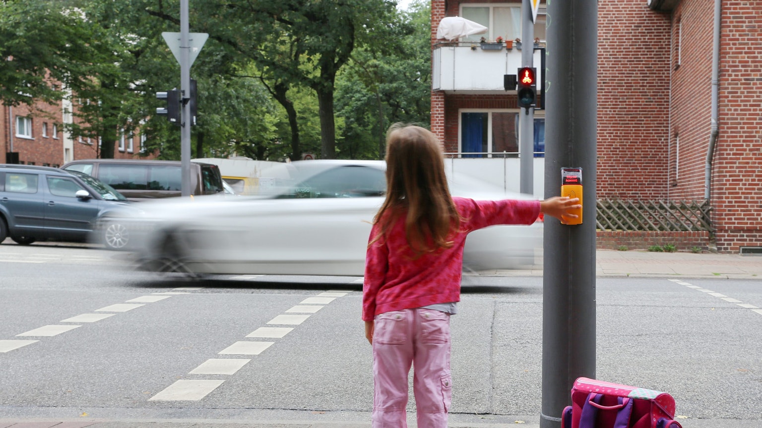 Ein rosa gekleidetes Mädchen steht mit Ranzen an einer Ampel, um die Straße zu überqueren.