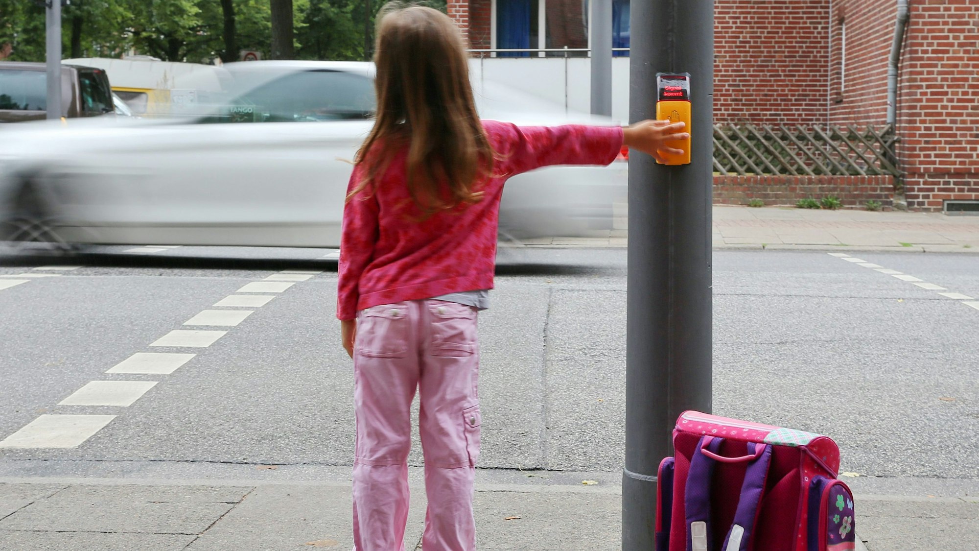 Ein rosa gekleidetes Mädchen steht mit Ranzen an einer Ampel, um die Straße zu überqueren.