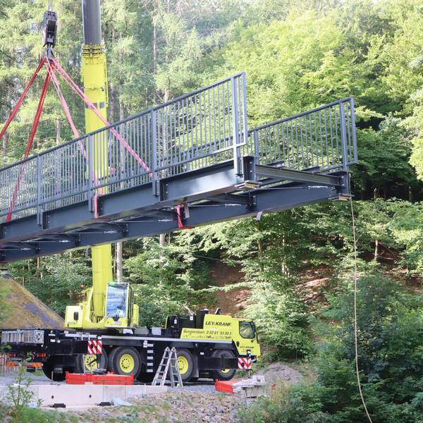 An einem Kran hängt eine graue, stählerne Brücke.