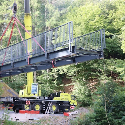 An einem Kran hängt eine graue, stählerne Brücke.