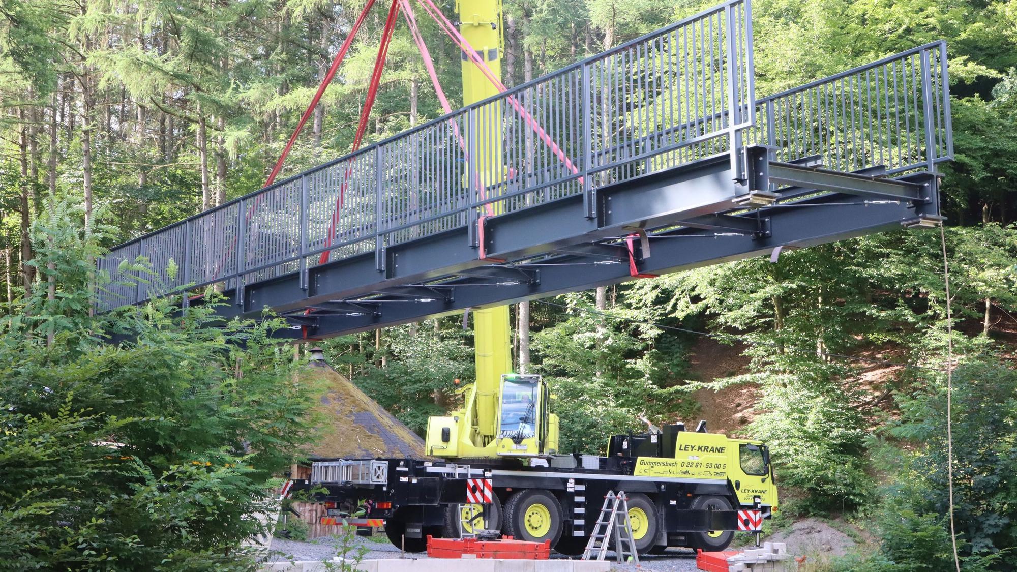 An einem Kran hängt eine graue, stählerne Brücke.
