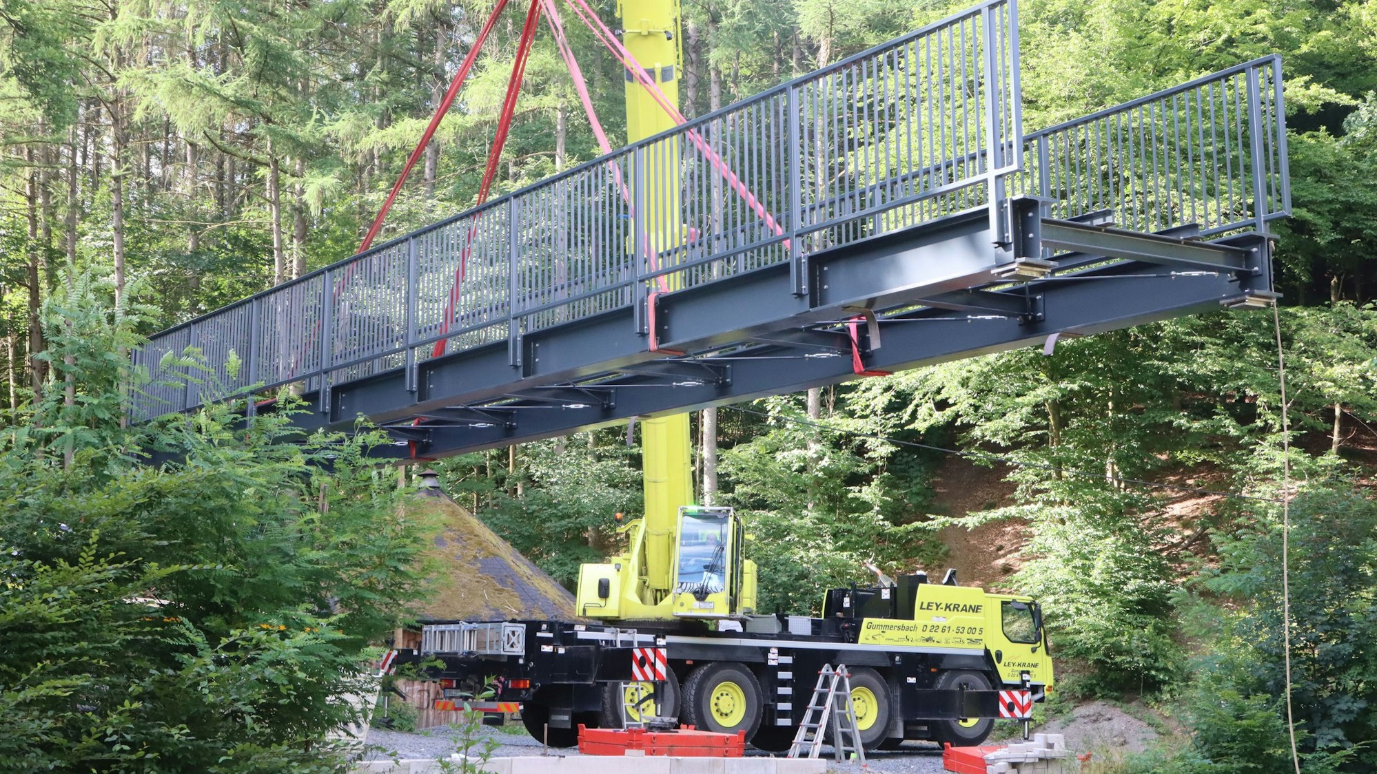 An einem Kran hängt eine graue, stählerne Brücke.