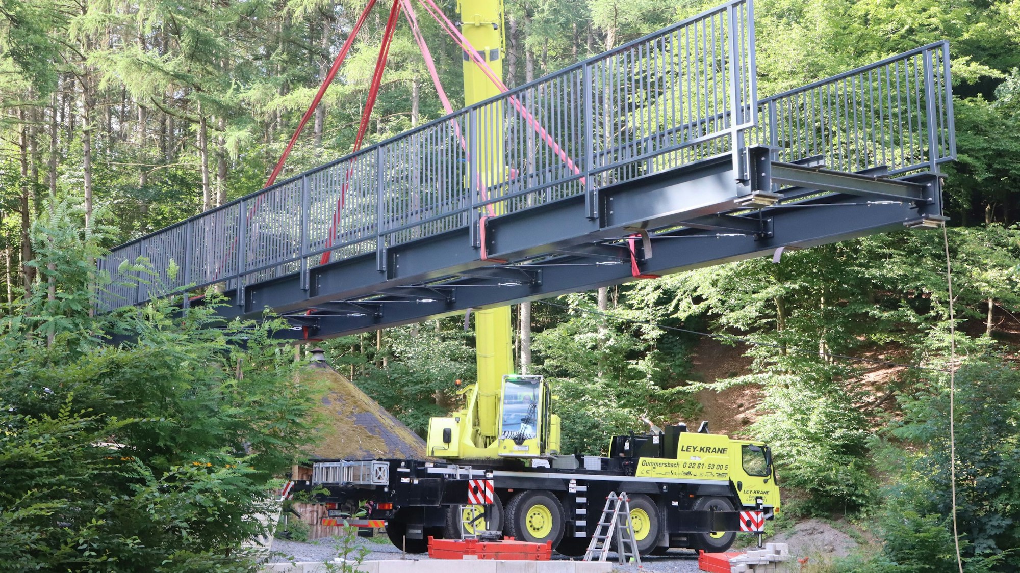 An einem Kran hängt eine graue, stählerne Brücke.