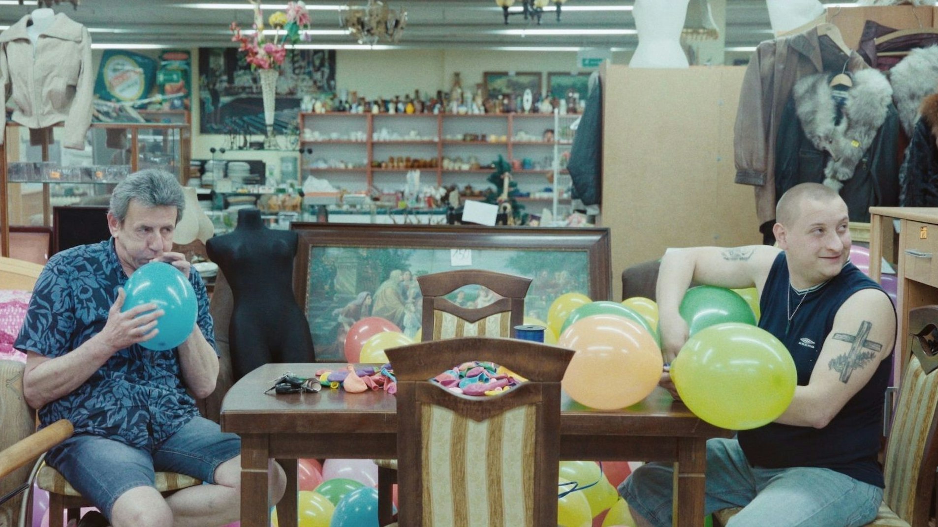 Zwei Männer sitzen in einem Trödelladen und blasen Luftballons auf.