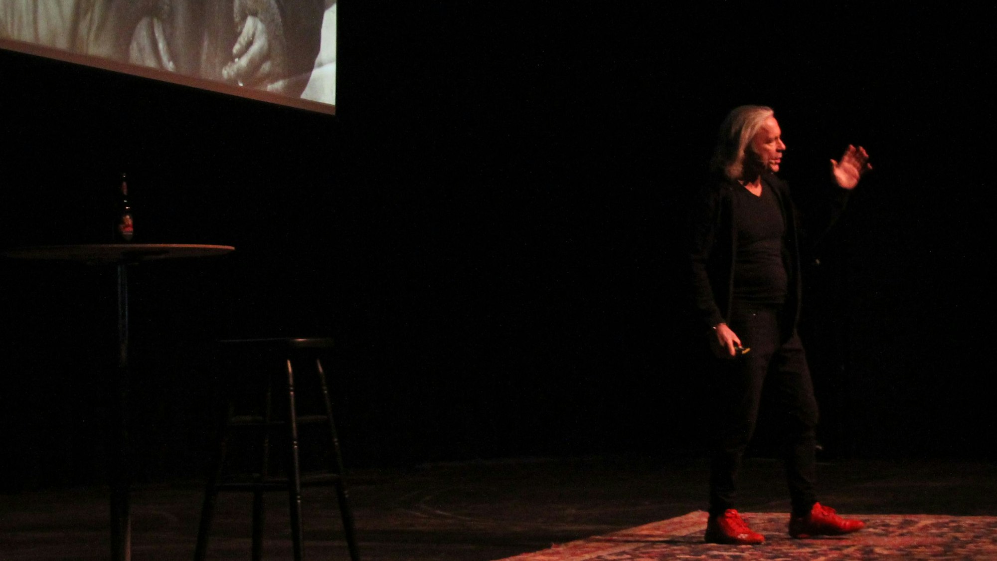 Iron-Maiden-Sänger Bruce Dickinson steht auf einer Bühne, auf der nur ein Stehtisch, ein Hocker und ein Teppich zu sehen sind. Im Hintergrund werden Fotos auf eine Leinwand projiziert.