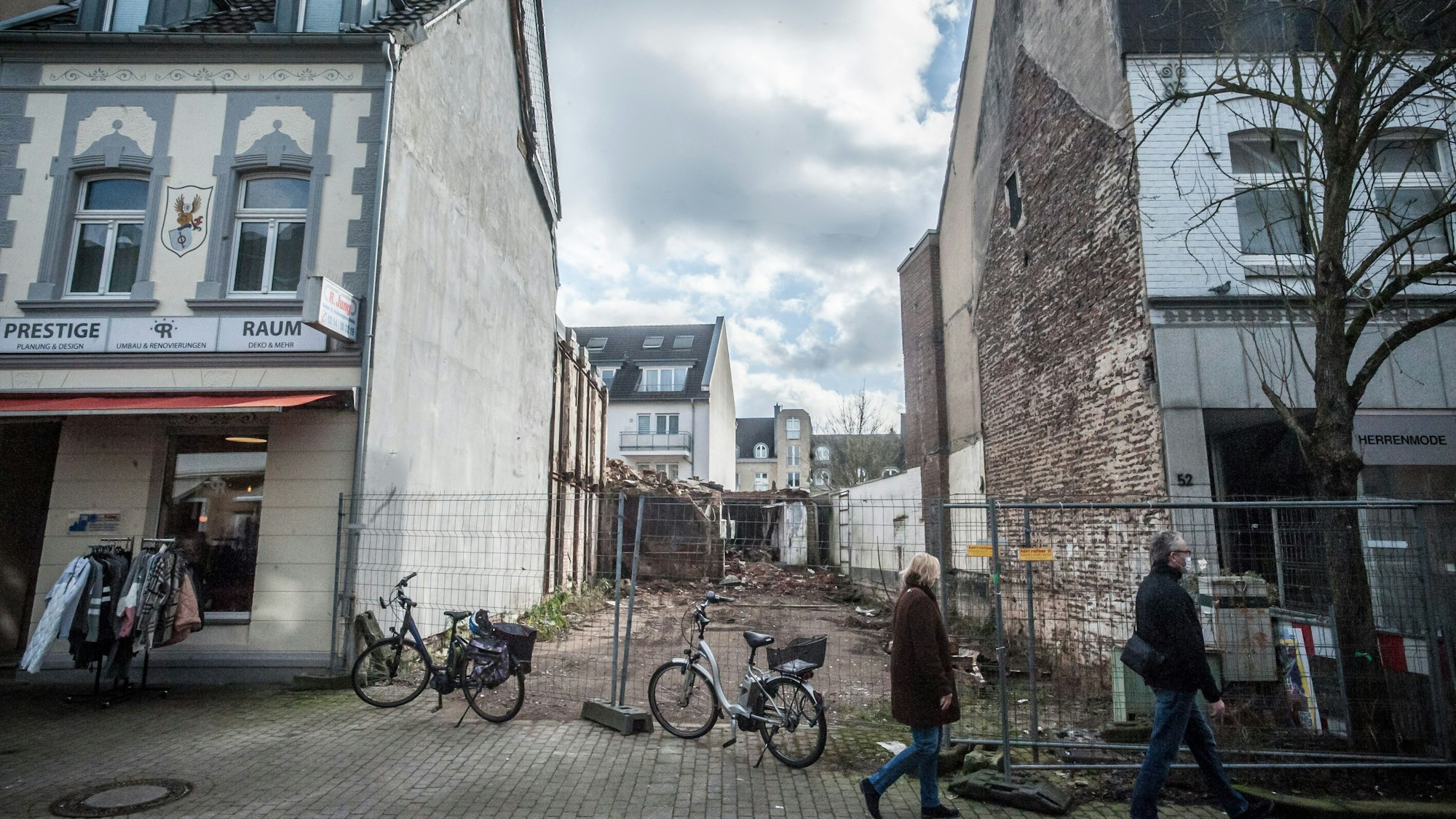 Blick auf die Brandruine in der Schlebuscher Fußgängerzone. In der Häuserzeile ist eine Lücke, davor stehen Bauzäune. Zwei Menschen gehen an den Zäunen entlang.