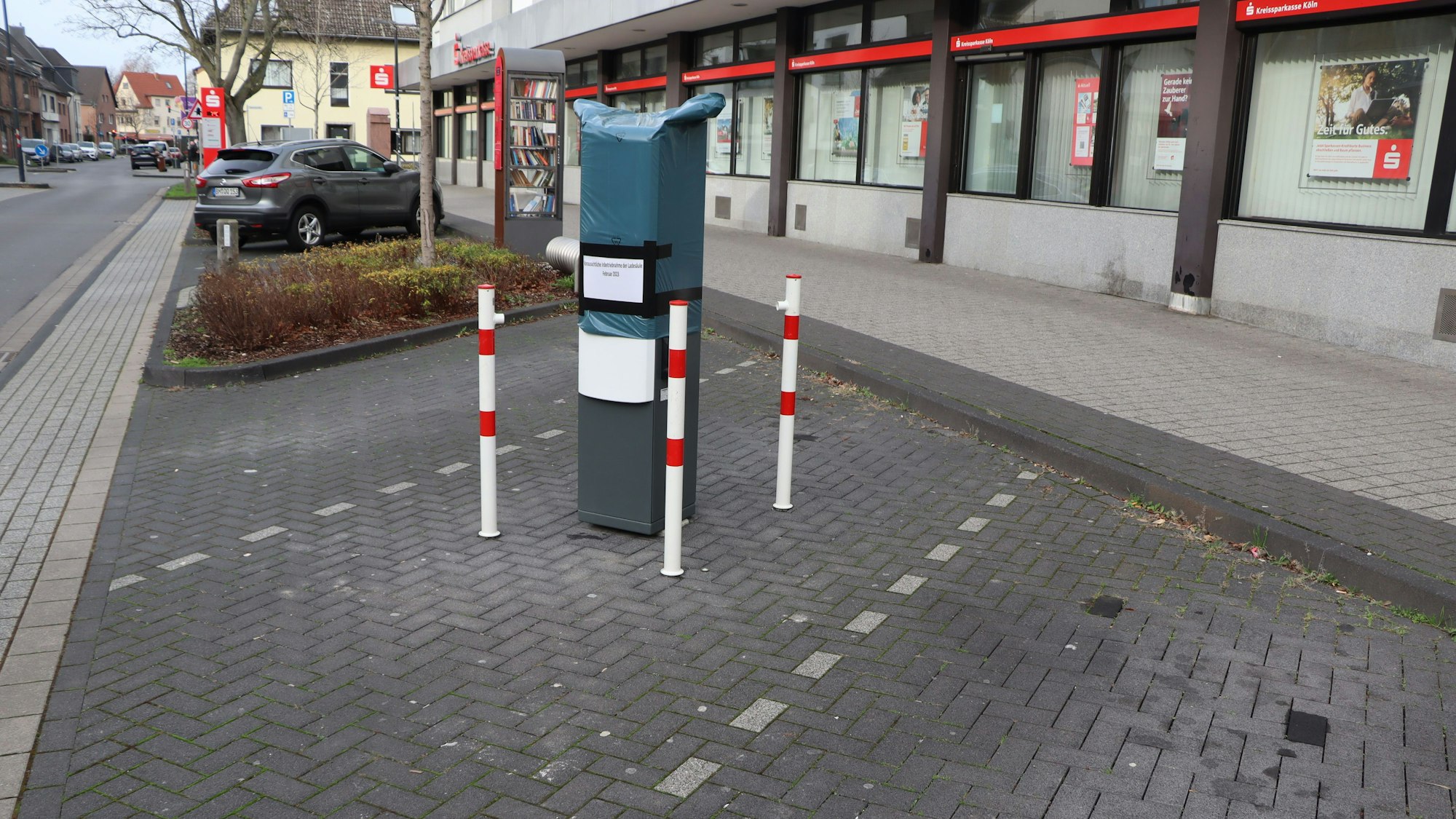 Mitten auf einer Parktasche wurde eine E-Ladesäule montiert. Drumherum stehen rot-weiße Poller.