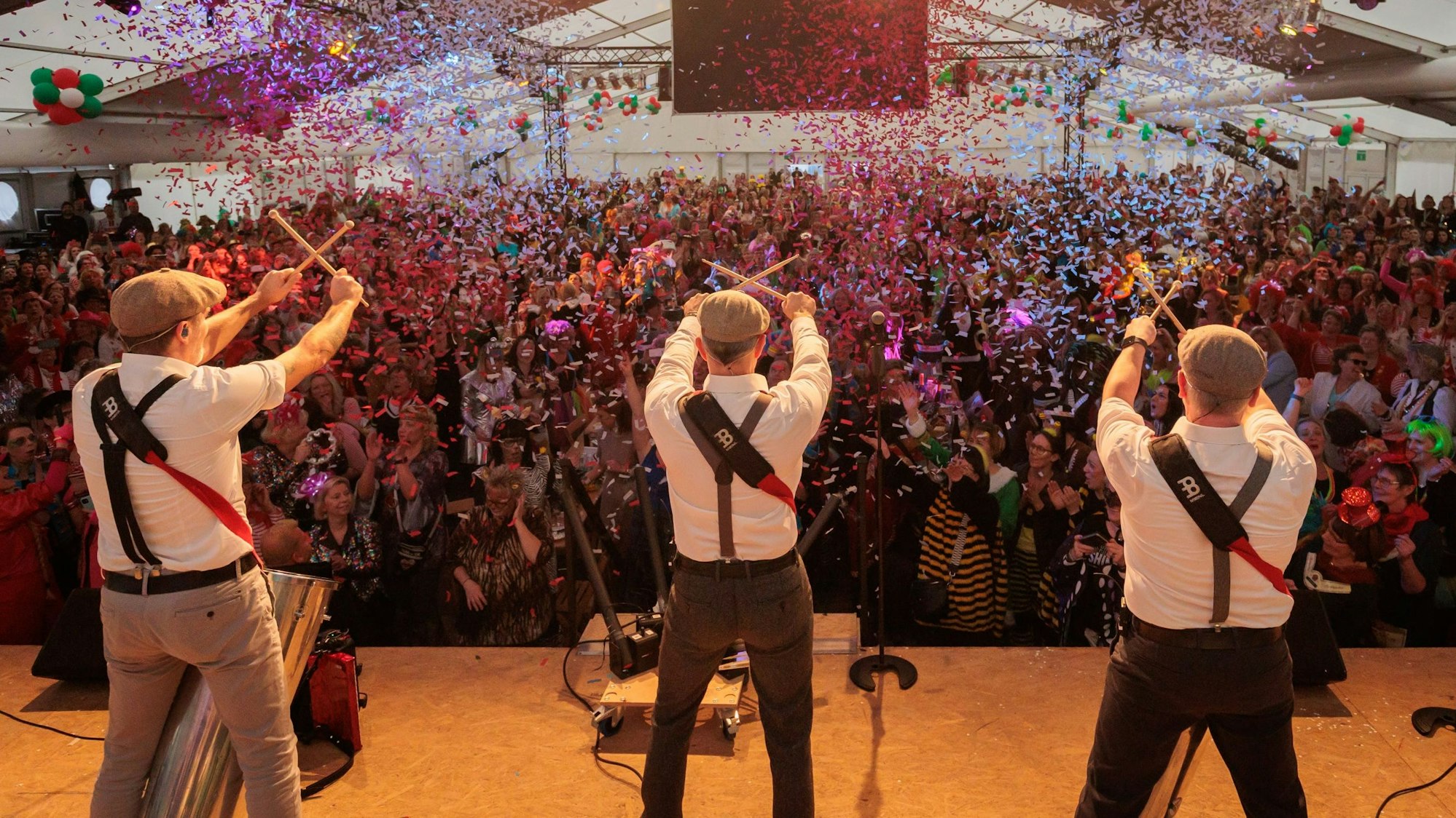 Auf einer Bühne stehen drei Musiker, die Drummer-Sticks in den Händen halten und diese vor den Gesichtern kreuzen. Vor der Bühne jubeln zahlreiche verkleidete Frauen. Von der Decke regnet Konfetti.