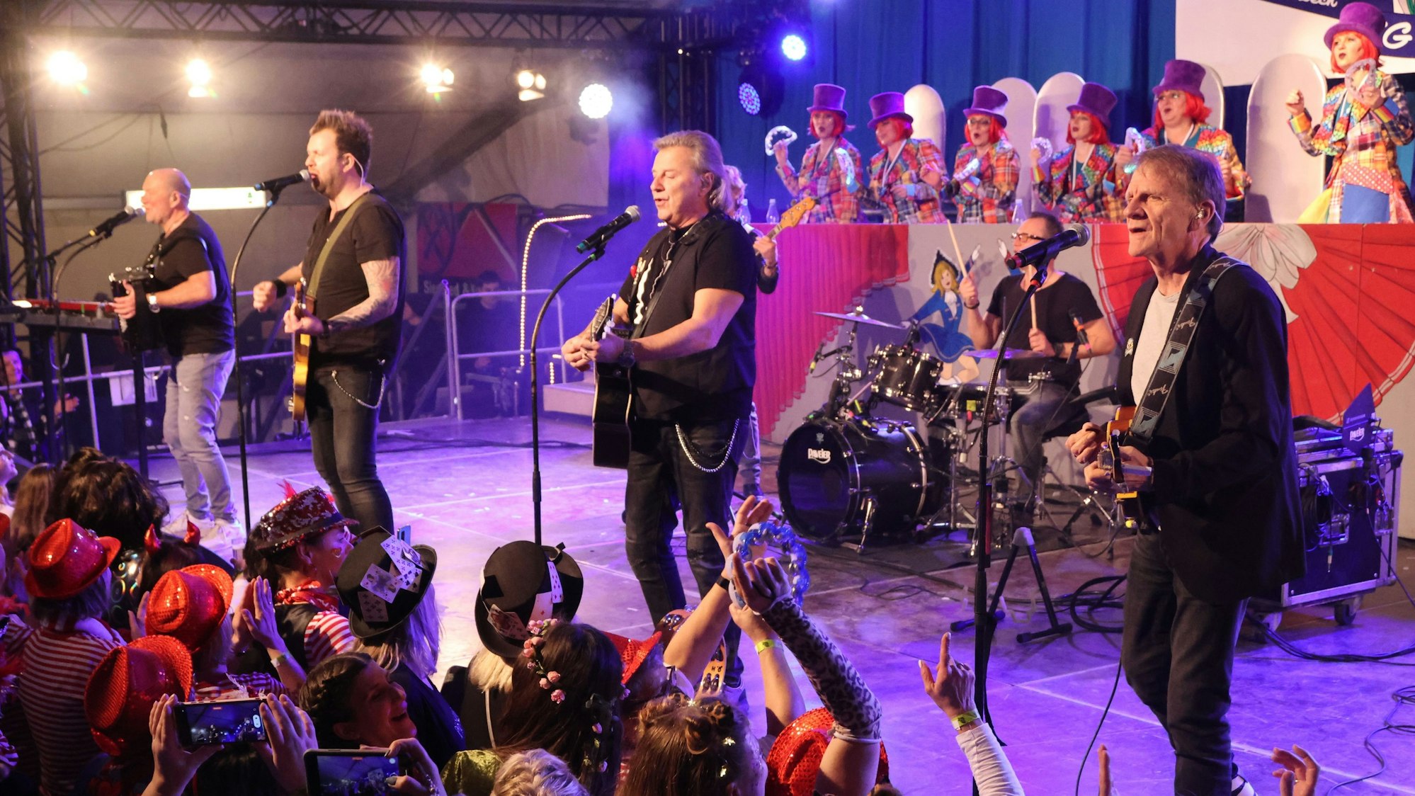 Vier Musiker stehen auf einer Bühne. Davor feiern verkleidete Damen Karneval.