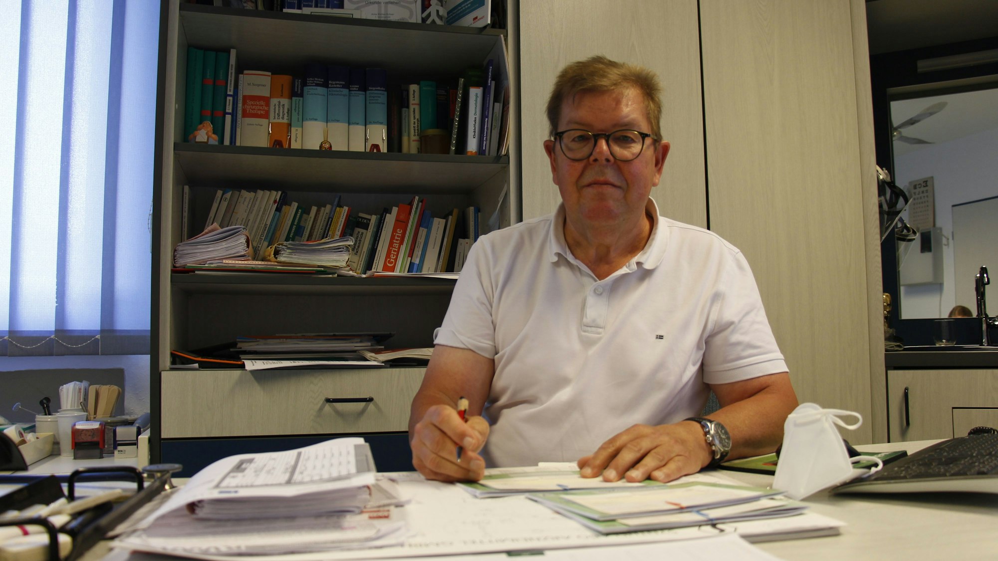 Pürschel sitzt an seinem Schreibtisch und guckt in die Kamera. In seinen Händen hält er einen Stift und einen Notizblock, hinter ihm steht ein Regal voller Bücher.