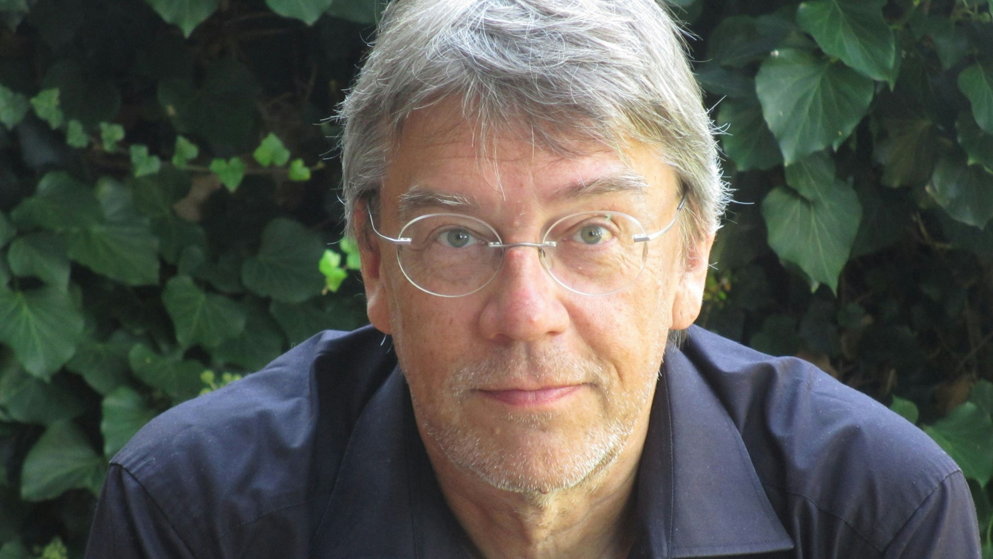 Thilo Winter, Autor von "Der Riss". Er trägt eine Brille und ein schwarzes Hemd. Im Hintergrund sind Blätter eines Busches.