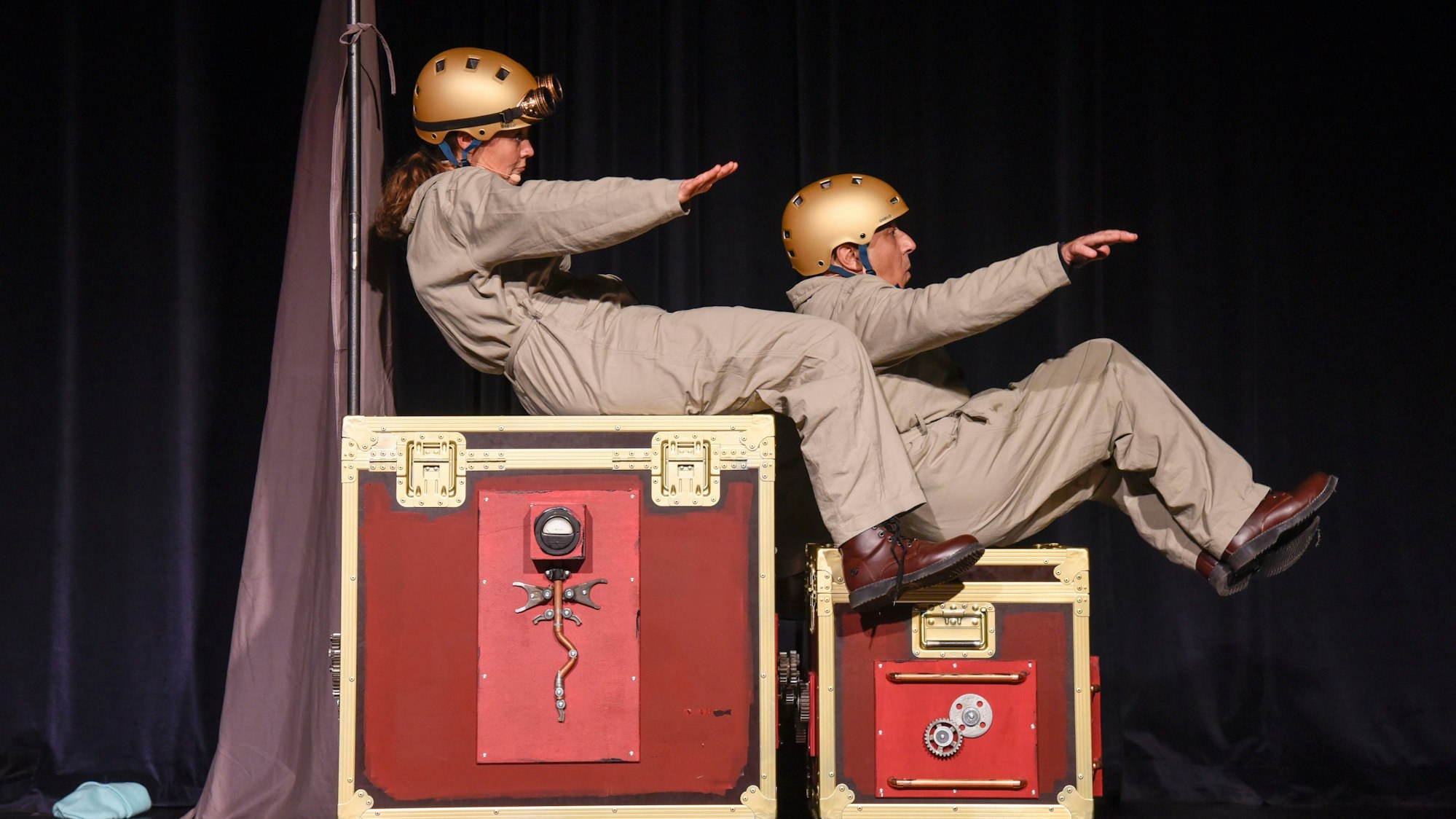 Zwei Schauspieler sitzen auf roten Kisten und machen Flugbewegungen.
