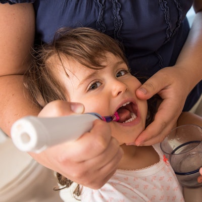 Die Hand eines Vaters putzt einem Kleinkind die Zähne mit einer elektrischen Zahnbürste.