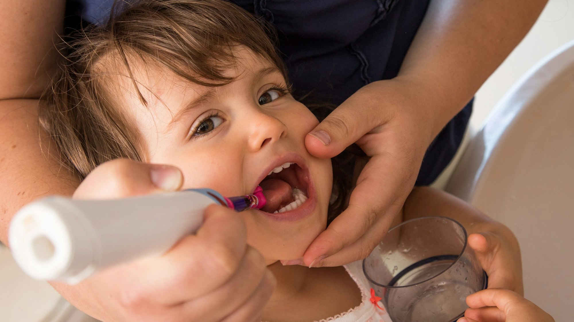 Die Hand eines Vaters putzt einem Kleinkind die Zähne mit einer elektrischen Zahnbürste.