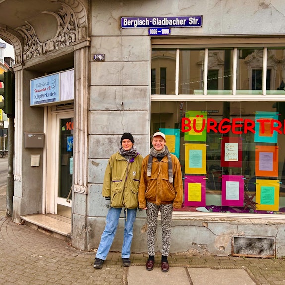 Vor einem Haus stehen eine Frau und ein Mann. In einem Fenster ist die Schriftzeile Bürgertreff zu sehen.