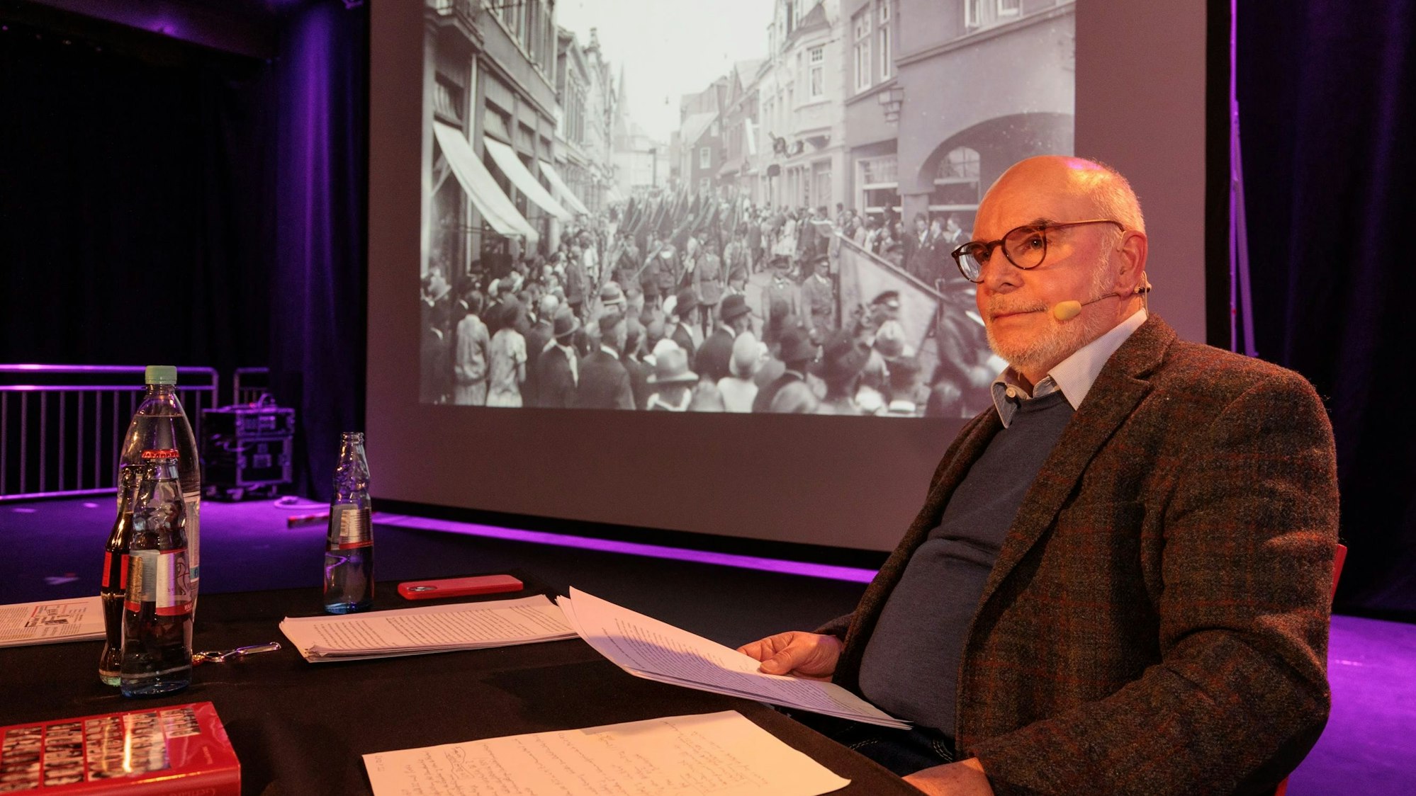 Ein älterer Mann sitzt an einem schwarzen Tisch auf einer Bühne und hält ein Blatt in der Hand. Hinter ihm ist ein historisches Bild auf einer Leinwand zu sehen, das Reichsbanner-Demonstrationszug zeigt.