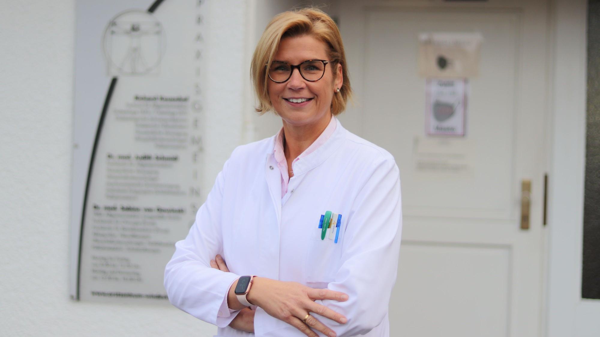 Judith Schmidt steht vor ihrem Praxiseingang. Sie trägt einen weißen Kittel, eine Brille und kinnlange, blonde Haare.