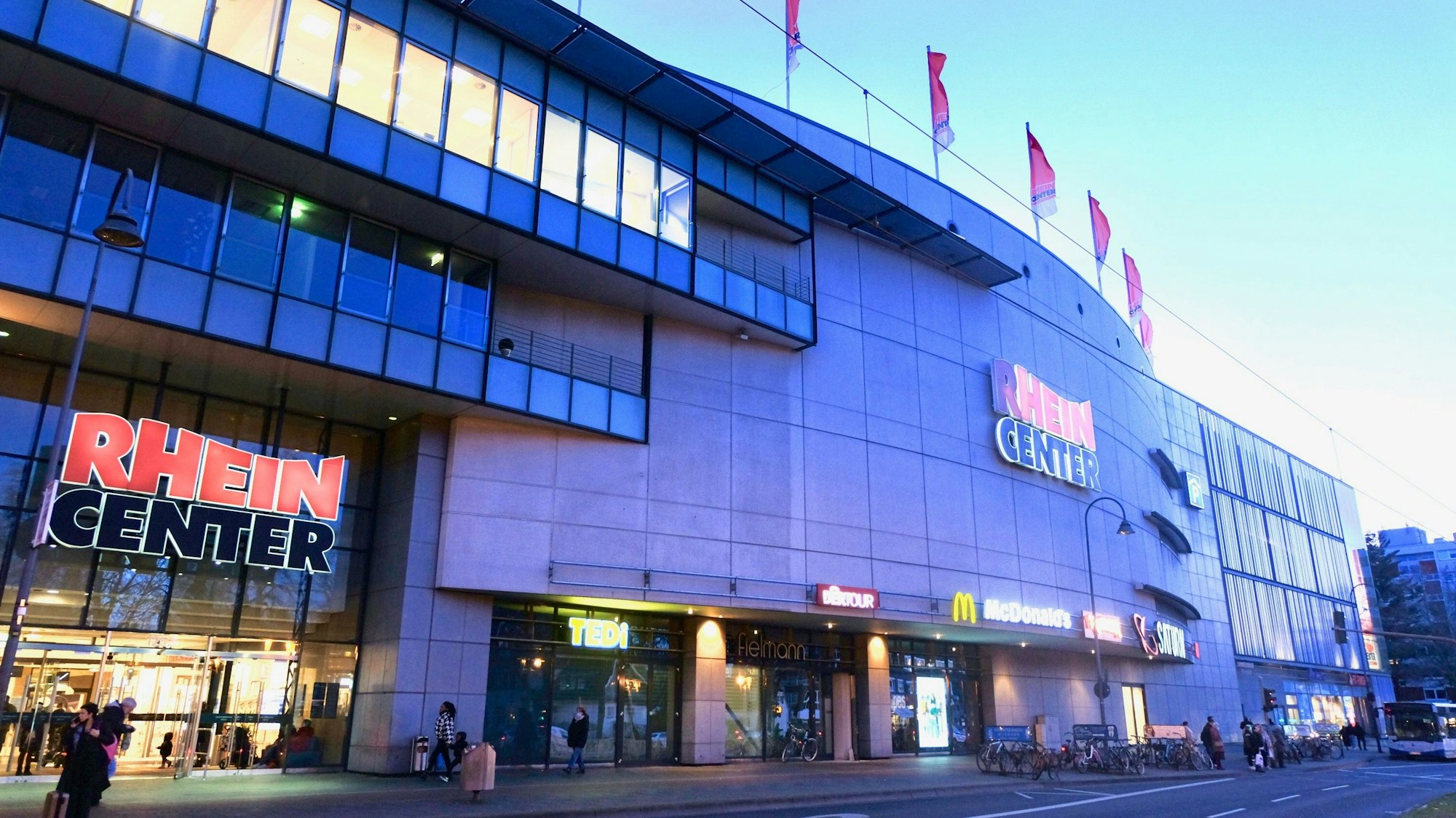 Ein mehrstöckiges Gebäude mit Fahnen auf dem Dach und dem Schriftzug Rhein-Center über Glas-Eingangstüren ist zu sehen.
