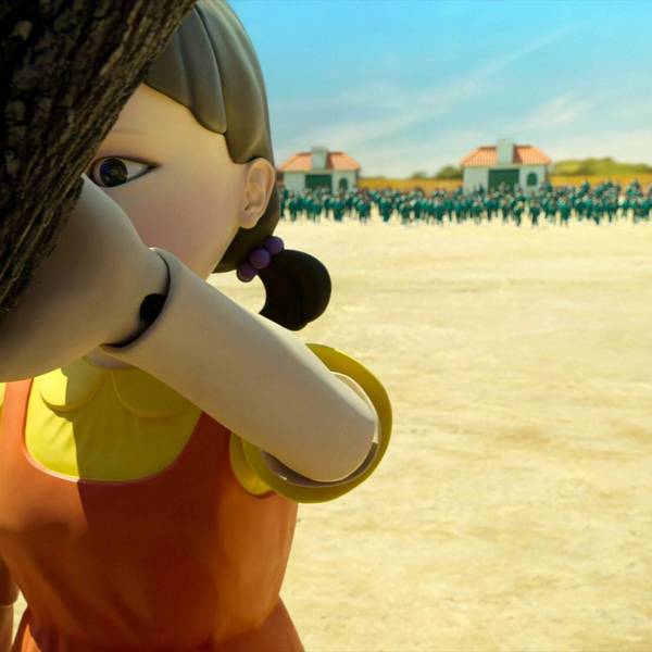 Eine Roboterpuppe lehnt in einer Szene aus der Netflix-Serie „Squid Game“ an einem Baum. Hinter ihr nähern sich die Teilnehmer des Spiels.