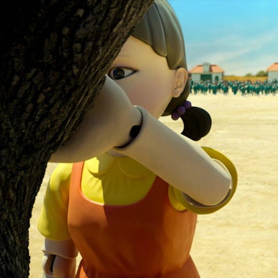 Eine Roboterpuppe lehnt in einer Szene aus der Netflix-Serie „Squid Game“ an einem Baum. Hinter ihr nähern sich die Teilnehmer des Spiels.