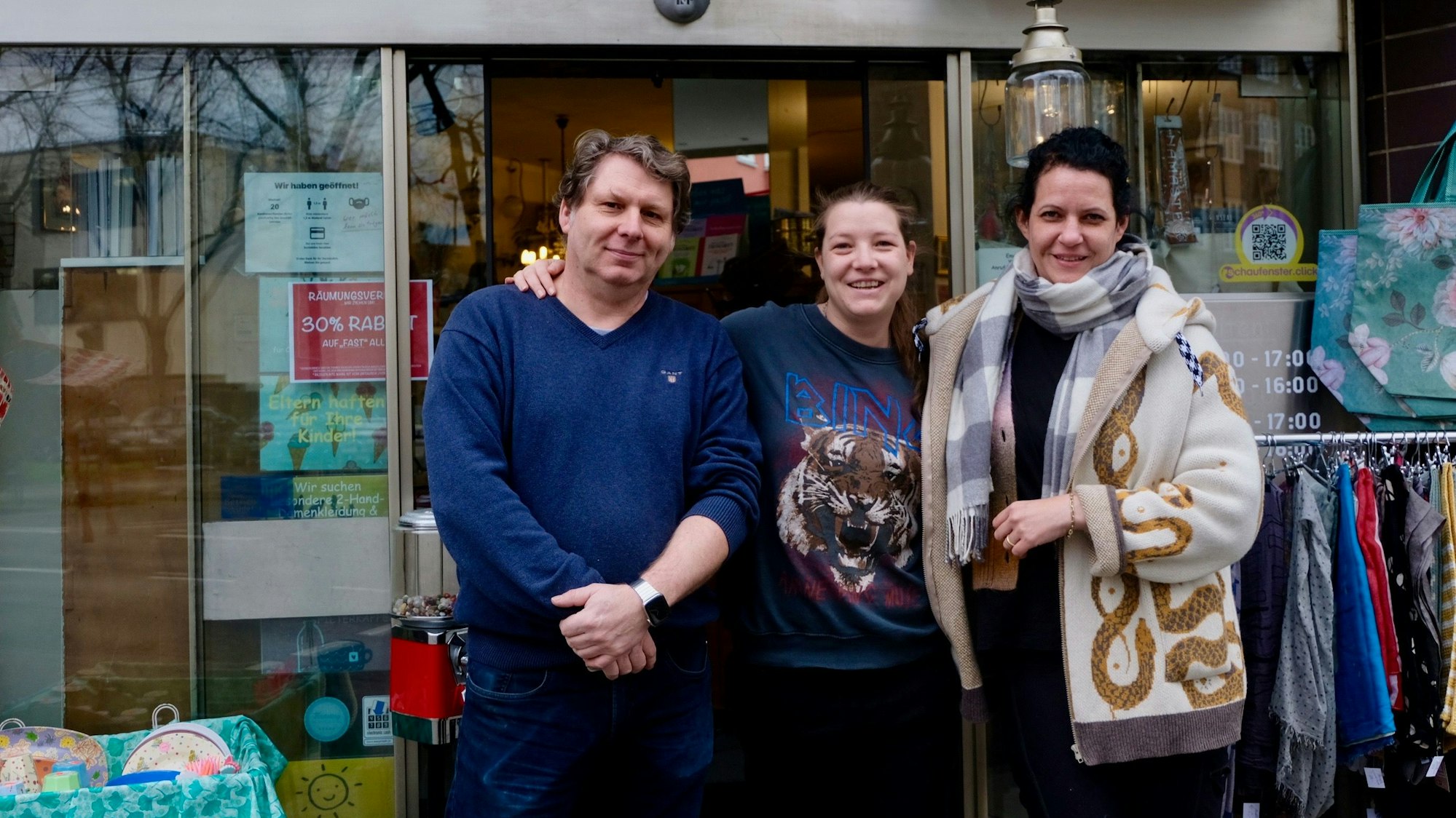 Vor einer Ladentür stehen ein Mann und zwei Frauen. Ein Ständer mit Kleidern ist zu sehen und bunte Taschen, die man in dem Laden kaufen kann.