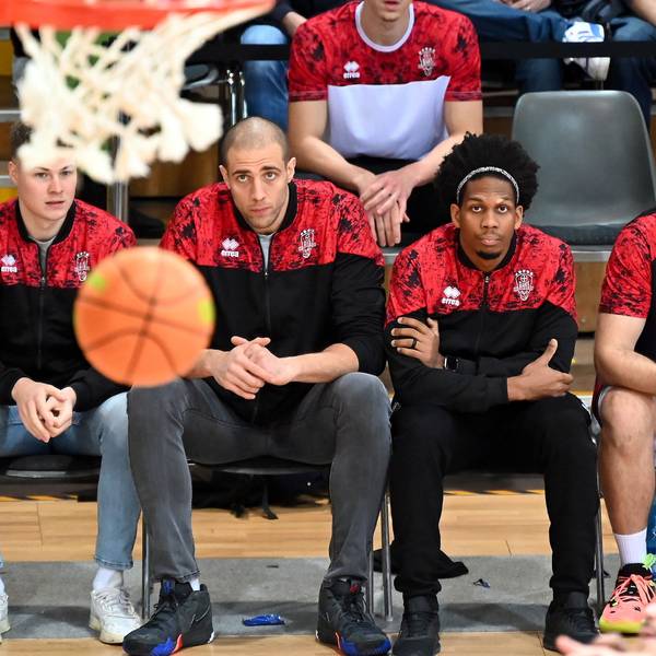 Drei Spieler der Bayer Giants sitzen auf der Bank. Mit ihrem Blick fokussieren sie einen Basketall, der in der Nähe des Korbs durch die Luft fliegt.