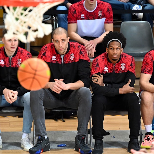 Drei Spieler der Bayer Giants sitzen auf der Bank. Mit ihrem Blick fokussieren sie einen Basketall, der in der Nähe des Korbs durch die Luft fliegt.