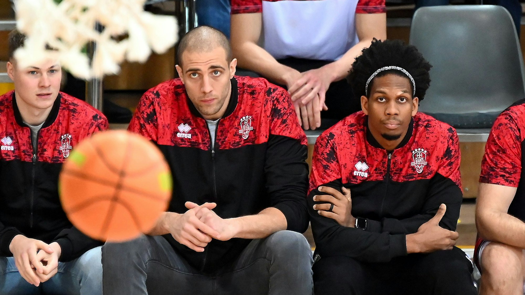 Drei Spieler der Bayer Giants sitzen auf der Bank. Mit ihrem Blick fokussieren sie einen Basketall, der in der Nähe des Korbs durch die Luft fliegt.