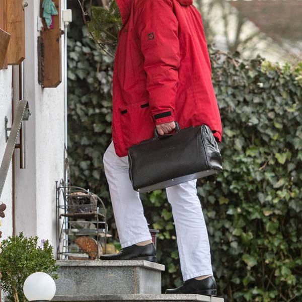 Eine Person in Arzthose und mit einer schwarzen Tasche steht vor dem Eingang eines alten, dörflichen Hauses.