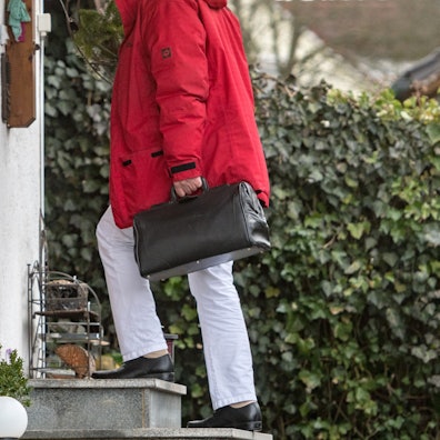 Eine Person in Arzthose und mit einer schwarzen Tasche steht vor dem Eingang eines alten, dörflichen Hauses.