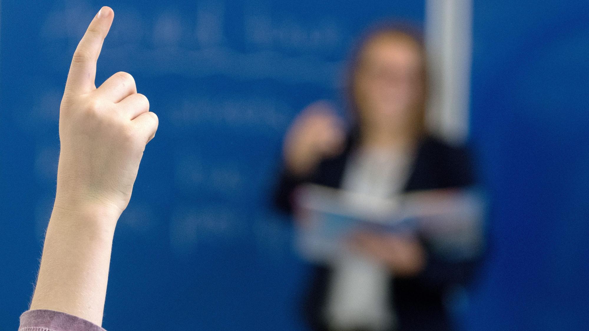 Das Bild zeigt einen Schüler, der sich meldet, dahinter eine Lehrerin an der Tafel. Der Lehrkräftemangel beschäftigt die Ständige Wissenschaftliche Kommission der Kultusministerkonferenz.
