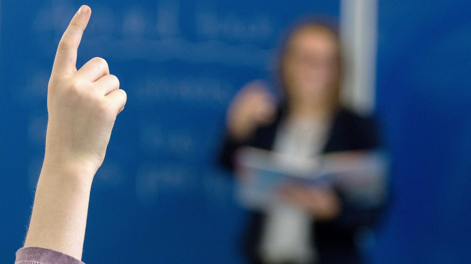 Das Bild zeigt einen Schüler, der sich meldet, dahinter eine Lehrerin an der Tafel. Der Lehrkräftemangel beschäftigt die Ständige Wissenschaftliche Kommission der Kultusministerkonferenz.