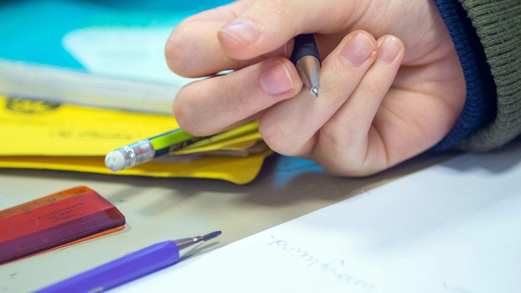 Eine Hand hält einen Stift in der Hand. Vor ihr liegen ein weißes Blatt, Stifte, Mappen und ein Lineal.