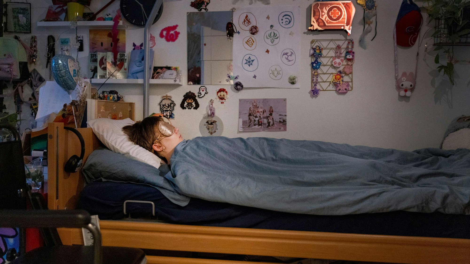Ed liegt in seinem Bett, er trägt eine Schlafmaske. Vor dem Bett steht ein Rollstuhl. An den Wänden kleben unzählige Bilder, Zeichnungen und Figuren.