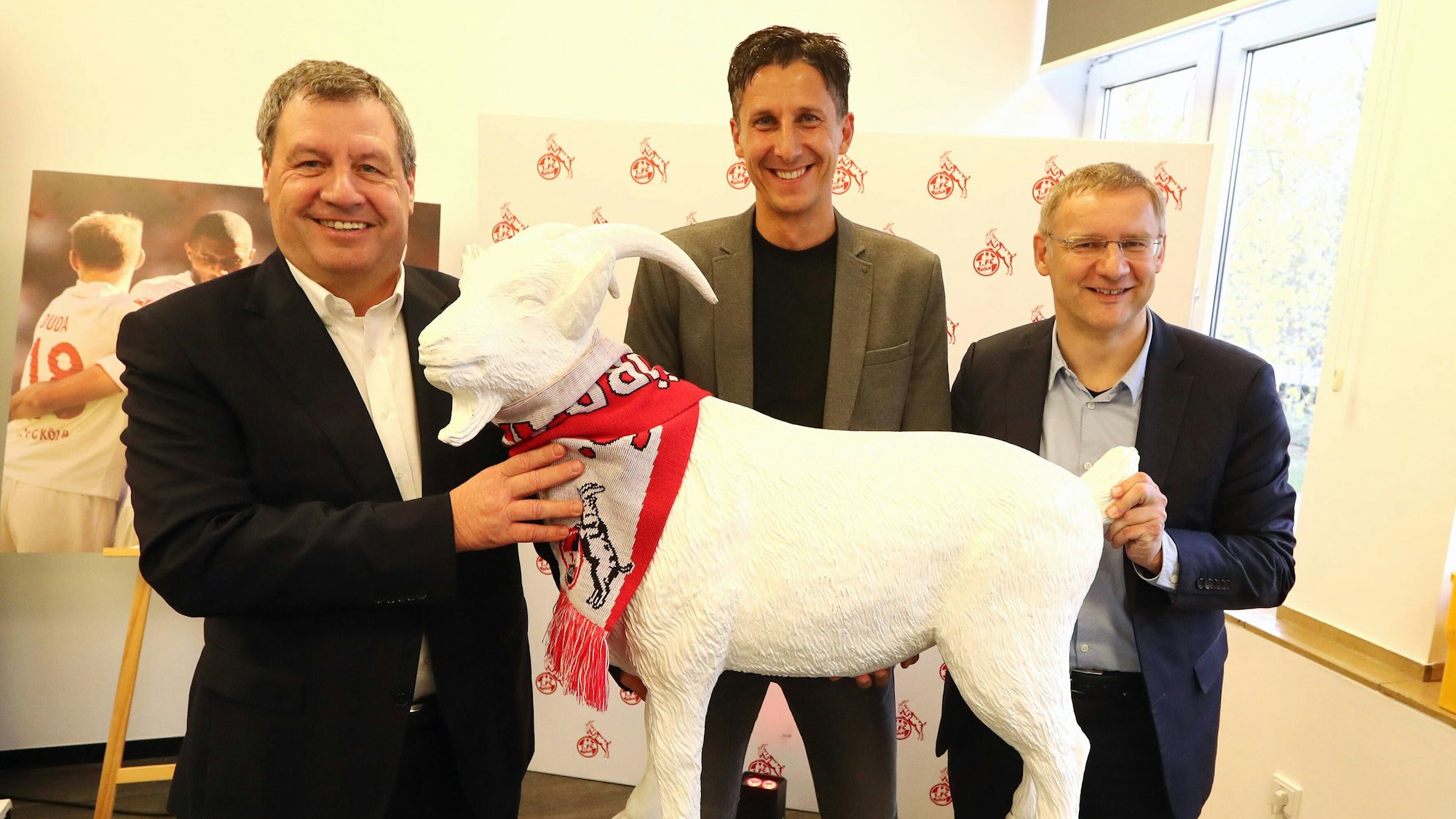 Präsident Werner Wolf und sein Vize Eckhard Sauren bei der Präsentation des Kölner Geschäftsführers Christian Keller. Die drei Herren halten gemeinsam einen Abguss des Kölner Geißbocks Hennes und lachen in die Kamera.