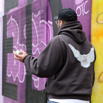 Der 22-jährige Amaru steht vor einer mit bunten Graffiti besprühten Mauer und hält eine Spraydose in der Hand.