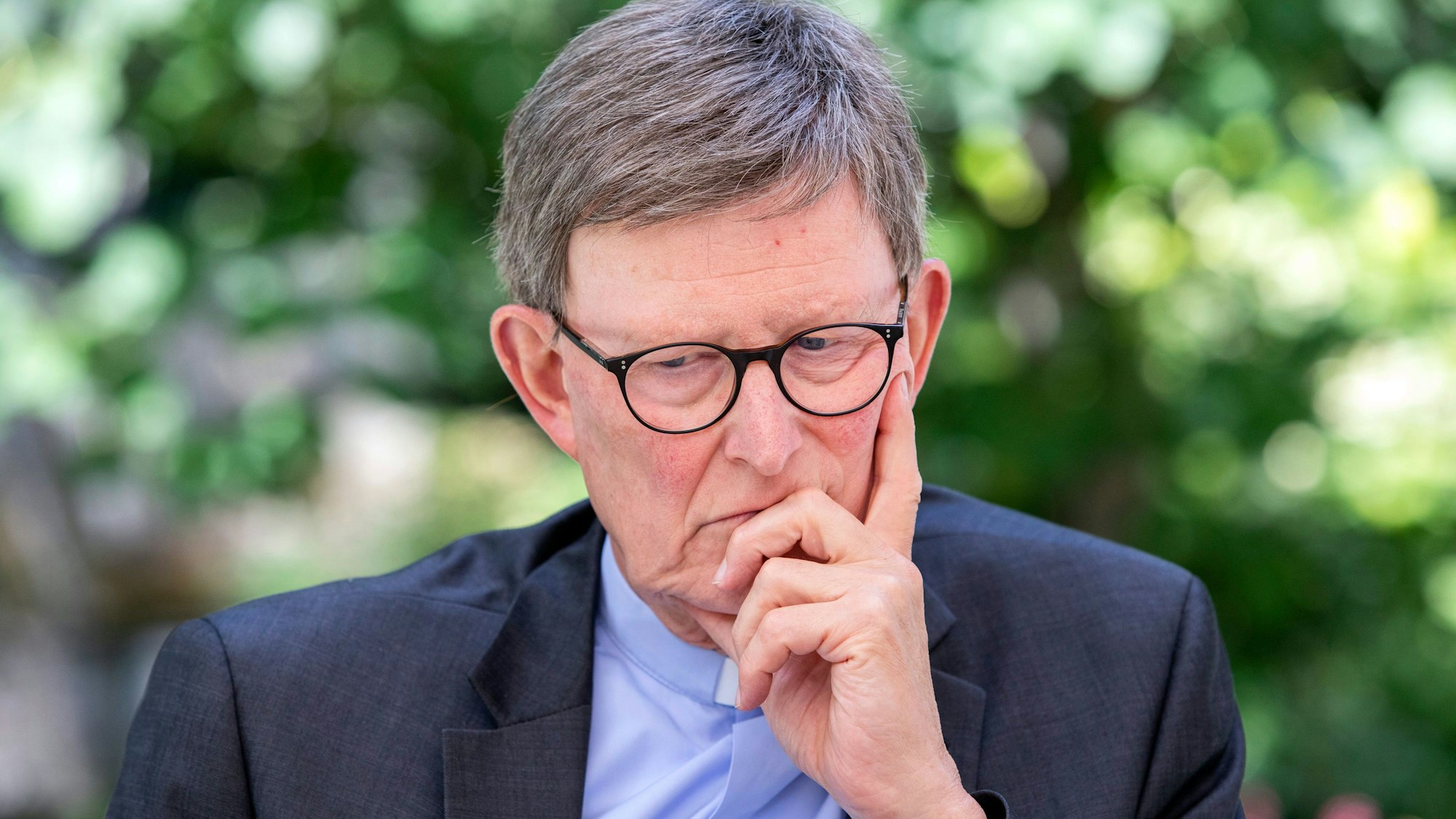 Erzbischof Rainer Maria Woelki sitzt mit nachdenklicher Geste vor einem grünen Garten.