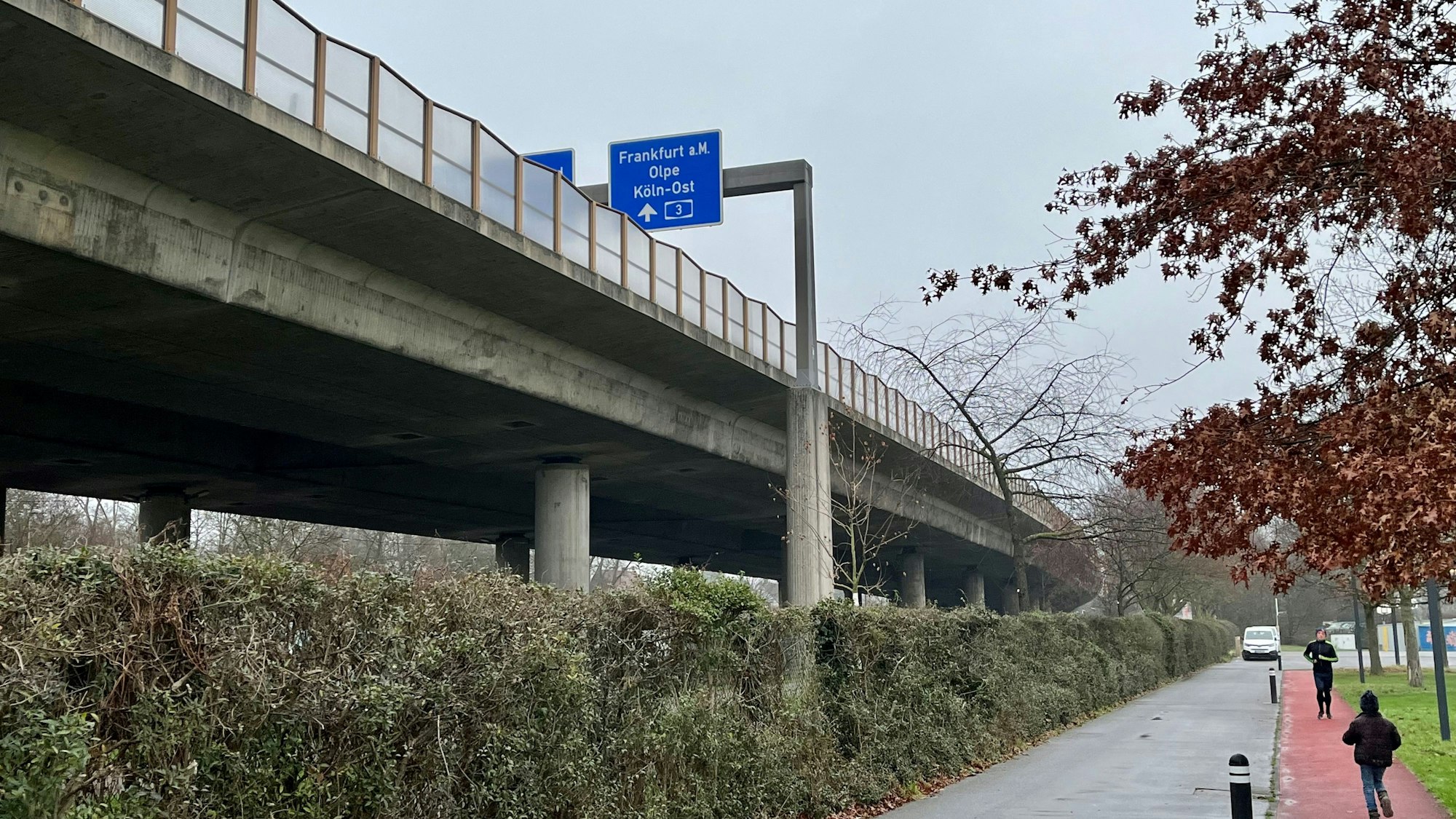 Zu sehen ist ein Gehweg entlang der Stelze der aufgebockten Autobahn 1 in Leverkusen, auf einem Autobahnschild ist zu lesen Richtung Frankfurt a. M. und Köln-Ost. Ein Mann joggt dort entlang.