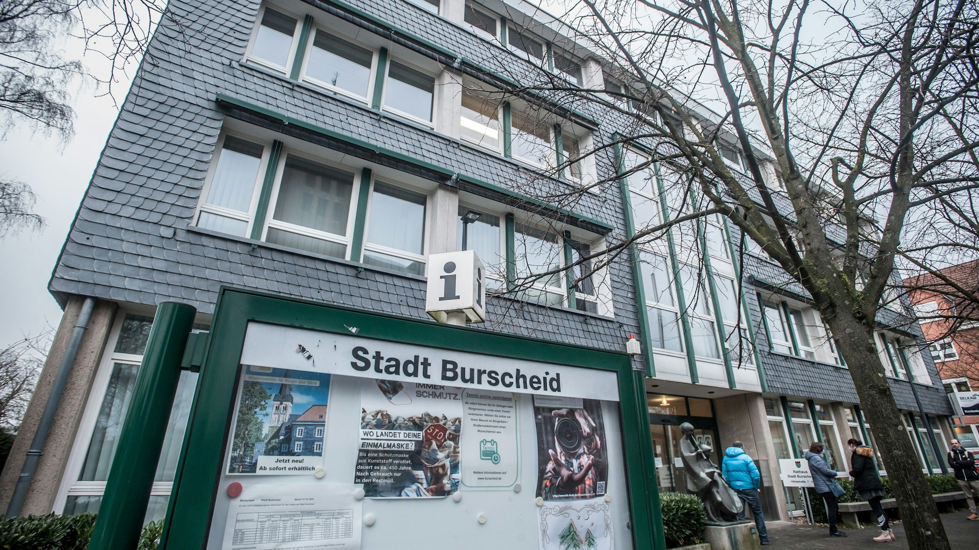Eine grün gerahmte Info-Vitrine mit der Aufschrift „Stadt Burscheid“ vor dem verschieferten Rathaus der Stadt.