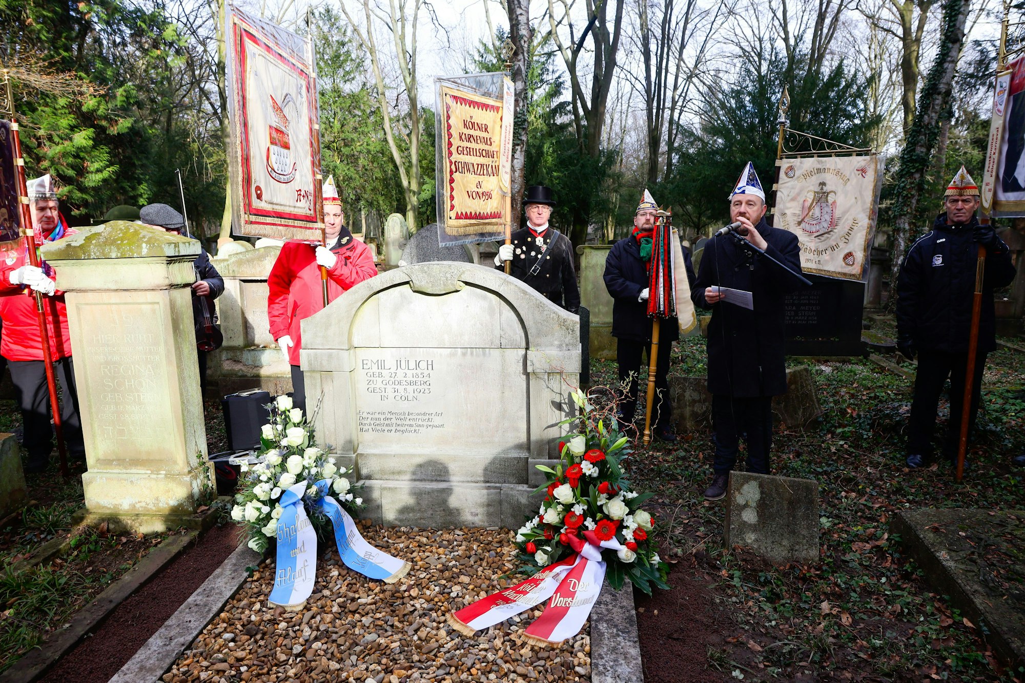 Aaron Knappstein spricht vor dem Grab von Emil Jülich. Neben ihm stehen Mitglider von Karnevalsvereinen, die ihre Fahnen tragen.